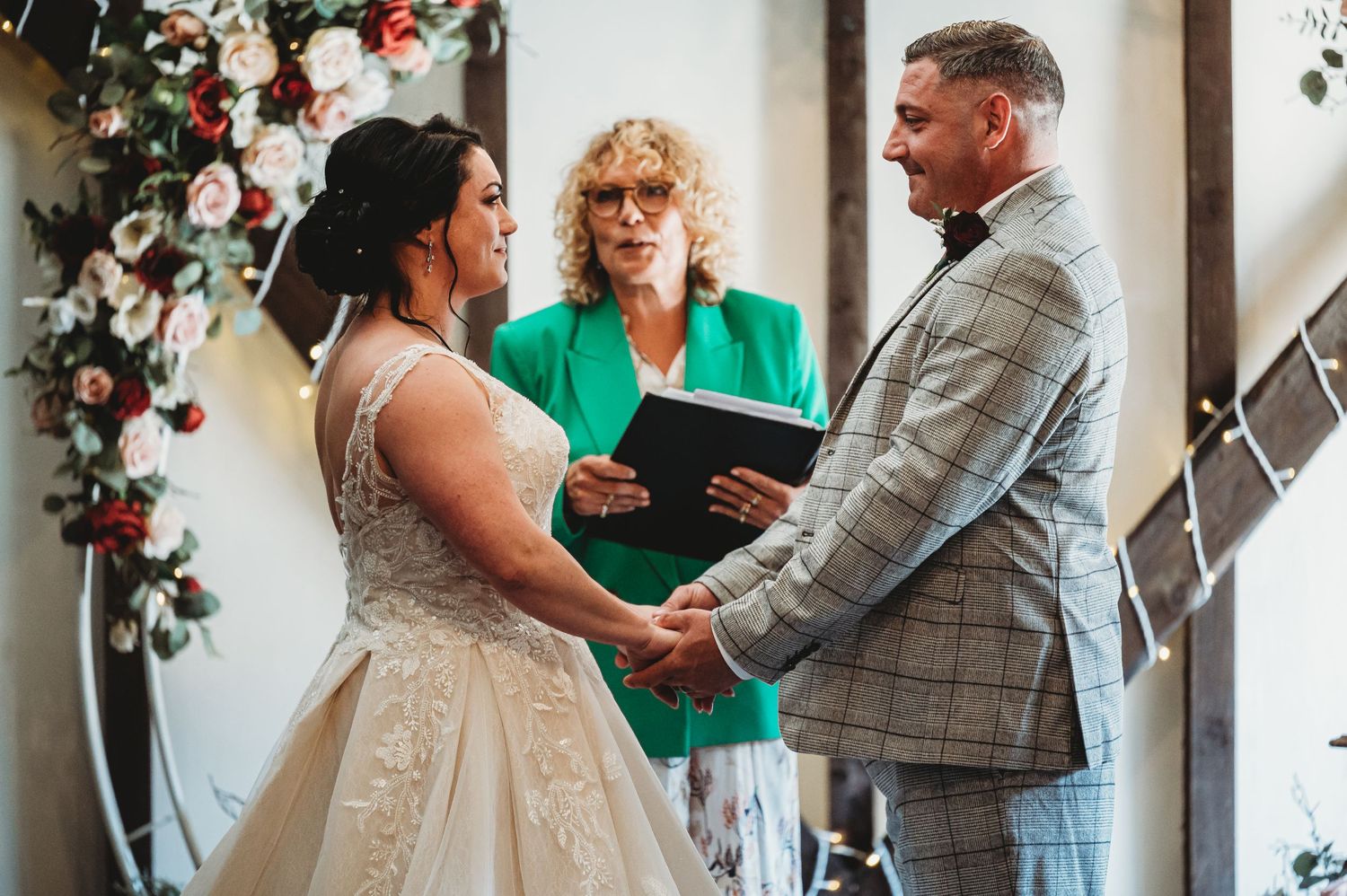 A couple holds hands during an intimate wedding ceremony with a person in teal officiating under floral arch decor.