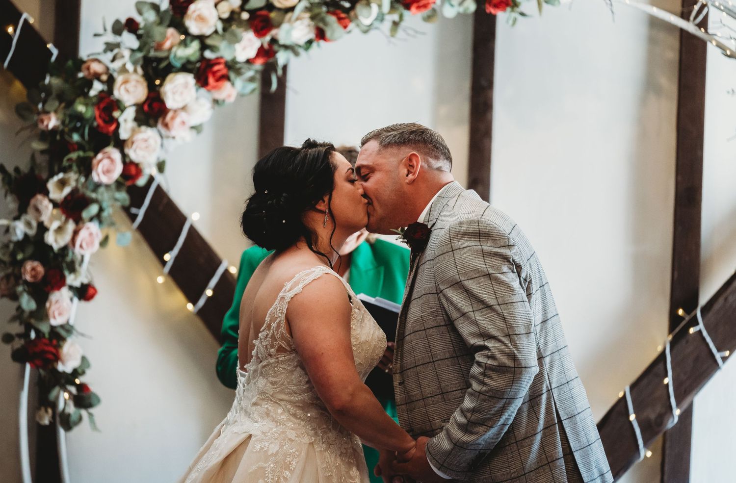 A couple shares a romantic kiss under an elegant archway decorated with red roses and white floral arrangements.
