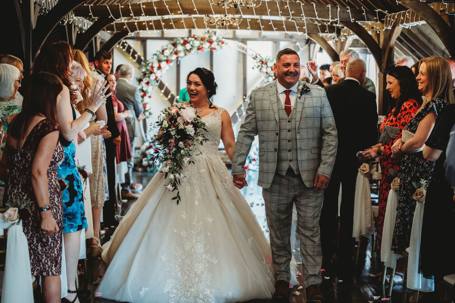 Wedding couple walks down rustic barn aisle while guests celebrate with sparklers under twinkle lights.