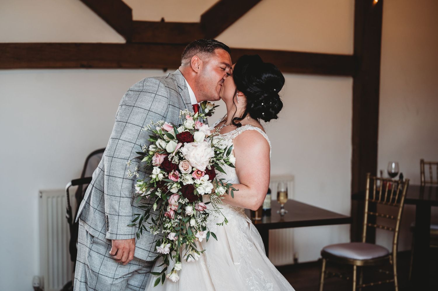 A romantic wedding couple share a kiss while holding a cascading bouquet of pink and white flowers in an indoor venue.