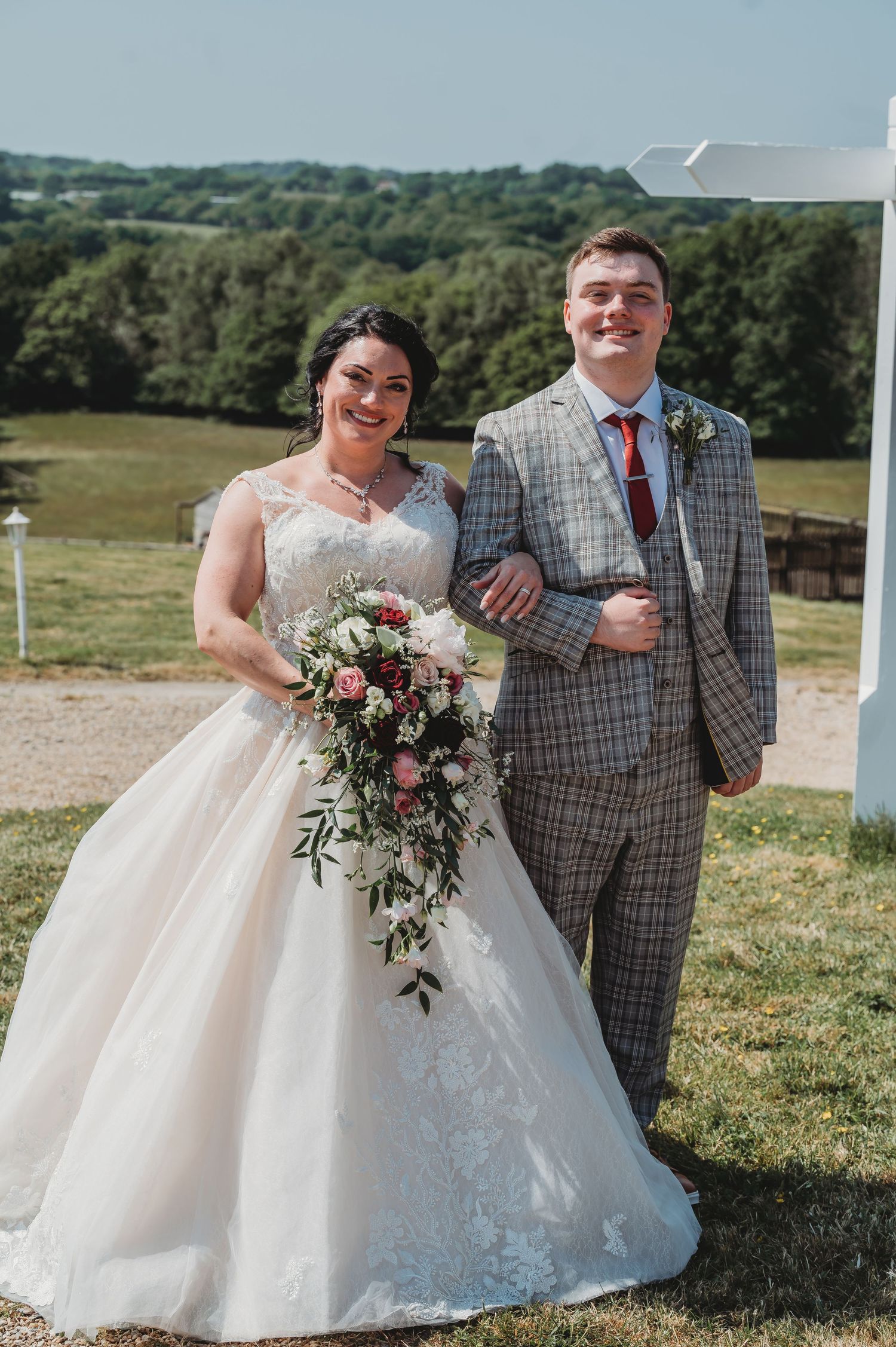 Happy couple poses at outdoor wedding venue with green hills and forest in background, featuring beautiful floral bouquet.