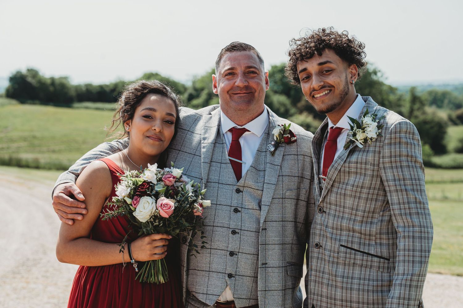 Three wedding guests pose together outdoors with bouquet of flowers on sunny summer day.