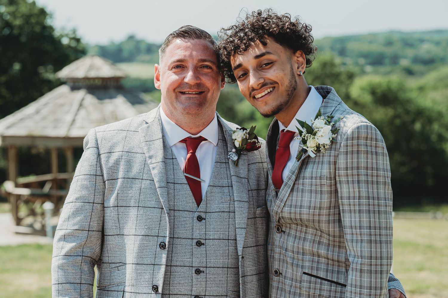 Two people in formal plaid suits and burgundy ties smile together outdoors with a gazebo in the background.