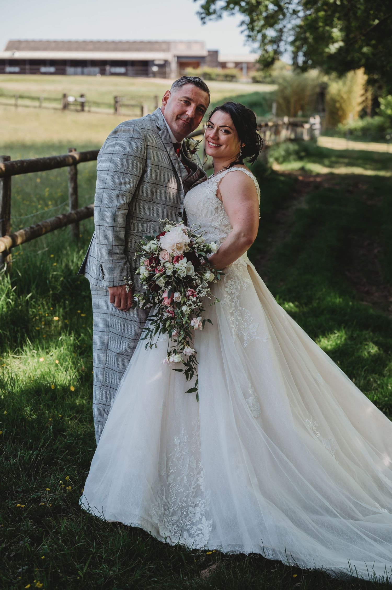 A couple poses together at their wedding celebrating in a lush outdoor setting with rustic wooden fences and fields.