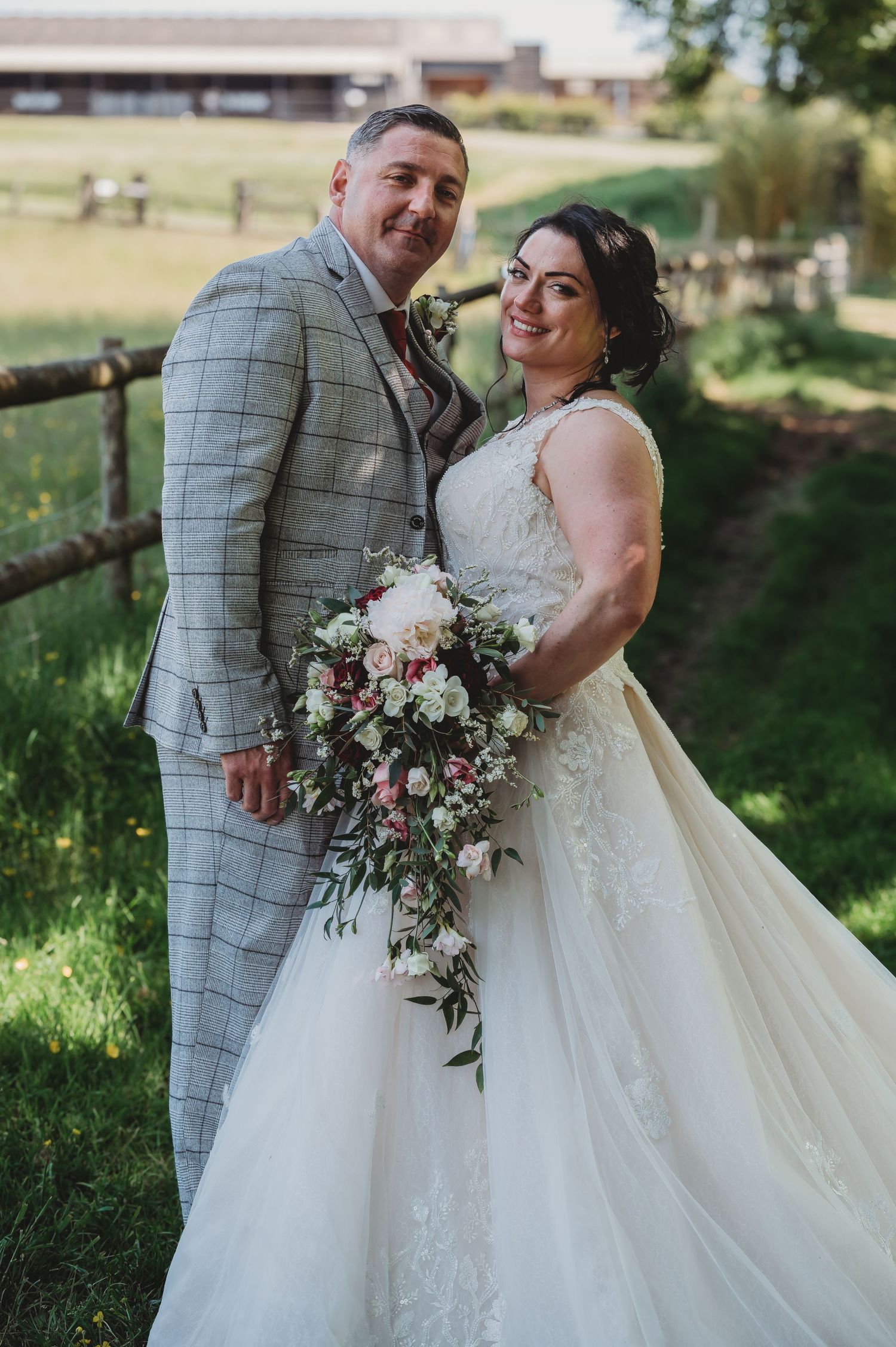 A couple poses outdoors at their rustic wedding, with the bride holding a cascading bouquet of blush and white flowers.