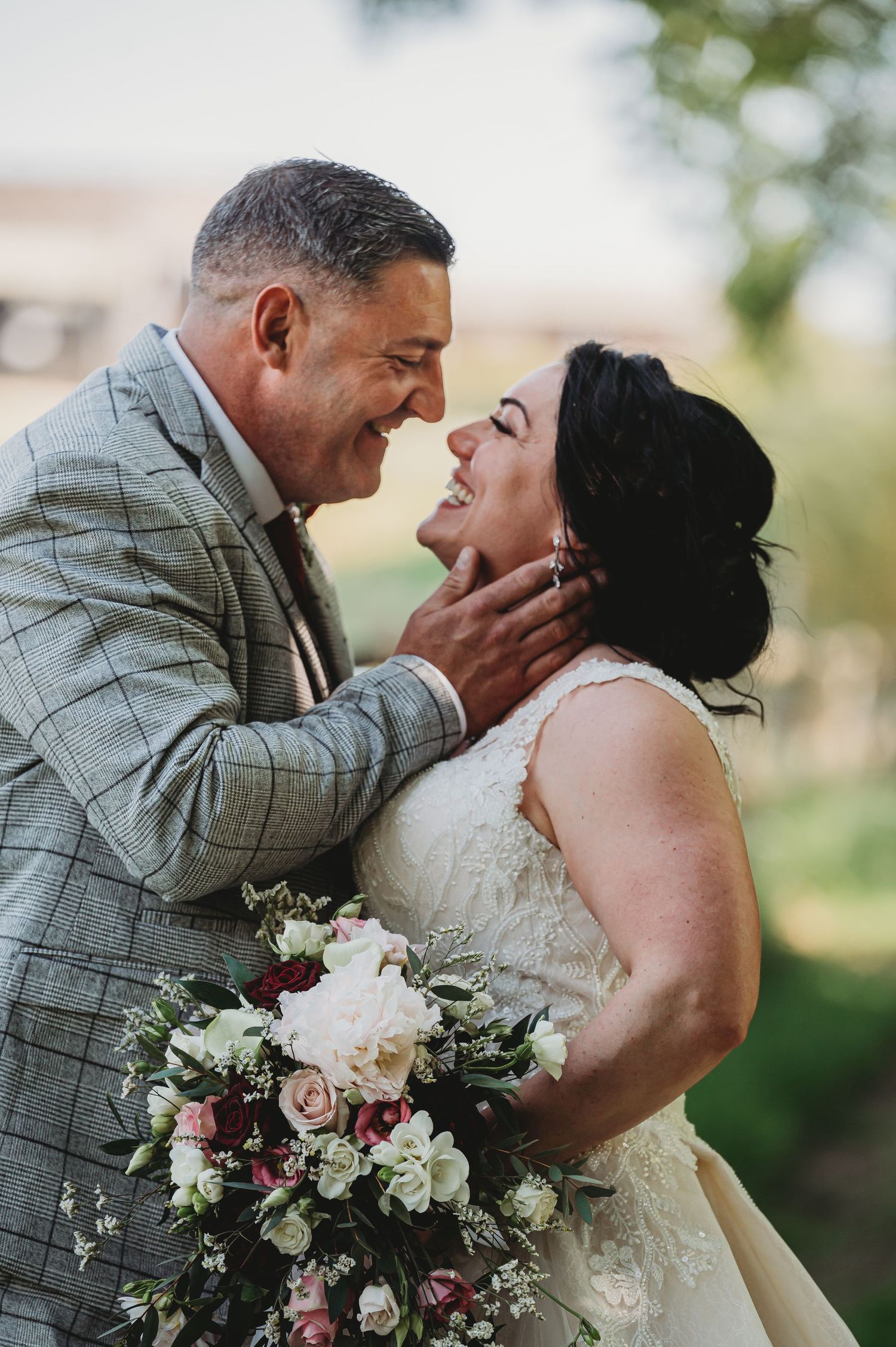 A romantic moment between newlyweds sharing laughter outdoors as the bride holds a beautiful floral bouquet.