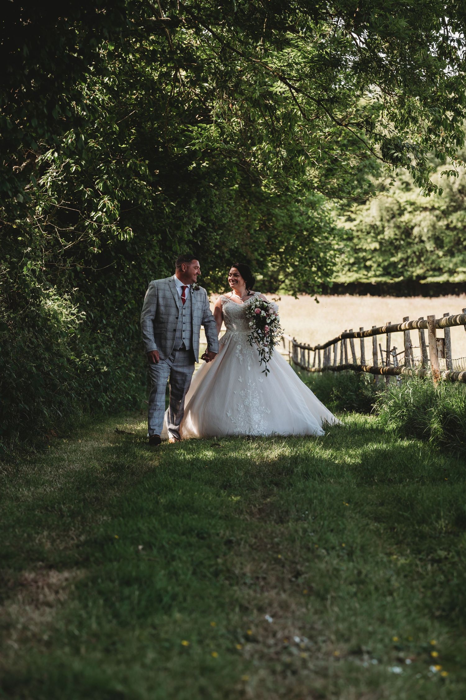 A romantic wedding couple walks through a sunlit garden path lined with lush greenery on their special day.