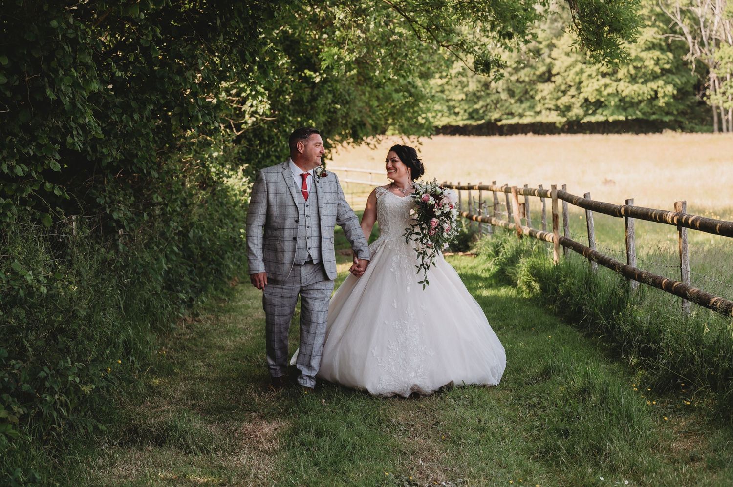 A newlywed couple walks hand in hand along a scenic country path lined with wooden fencing and lush greenery.
