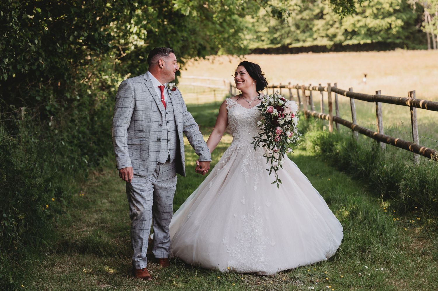 A couple in wedding attire walk hand in hand along a rustic fence-lined path during a summer countryside celebration.
