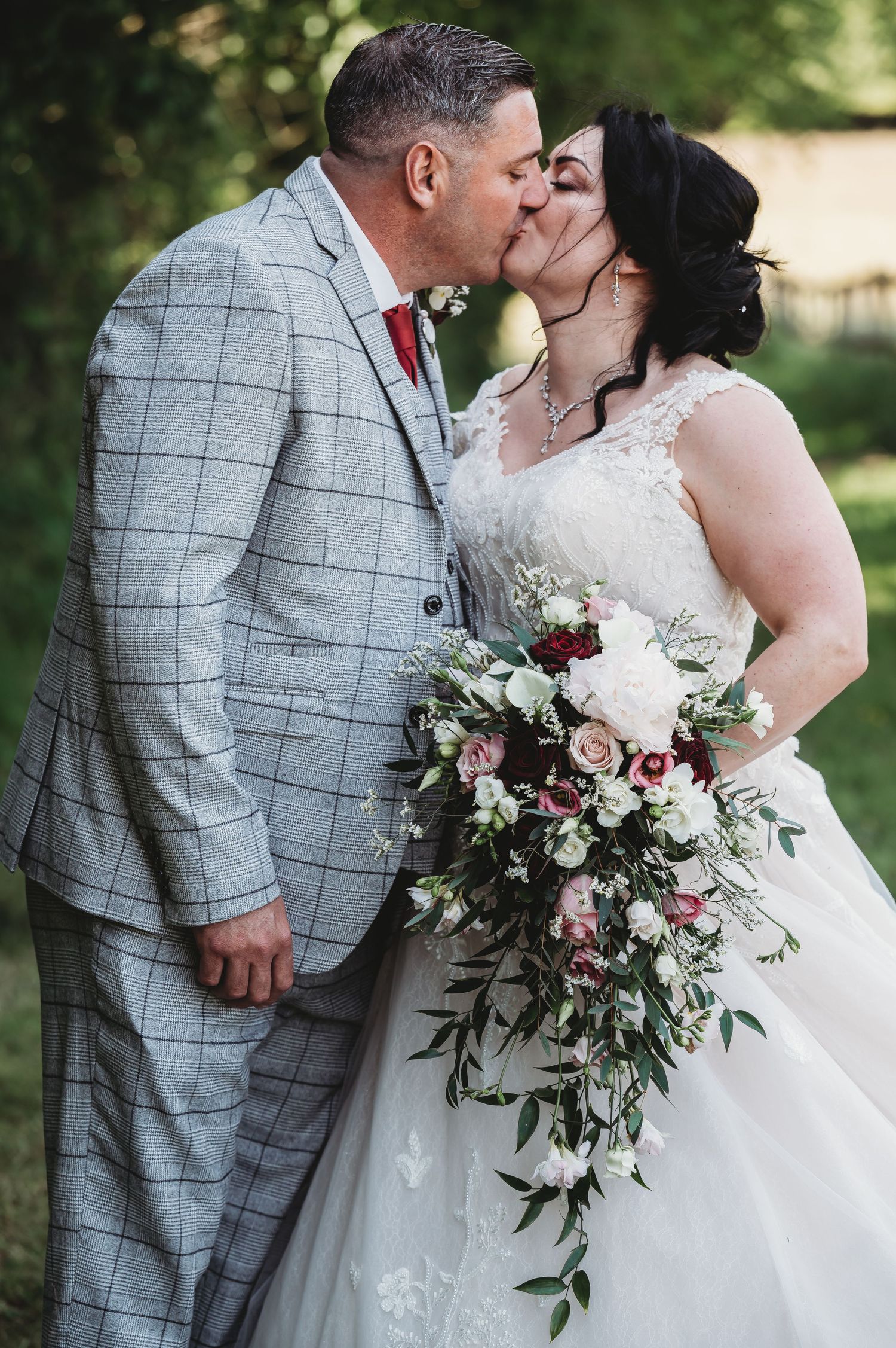 A romantic wedding scene outdoors with the bride holding a cascading bouquet of pink and white flowers with greenery.