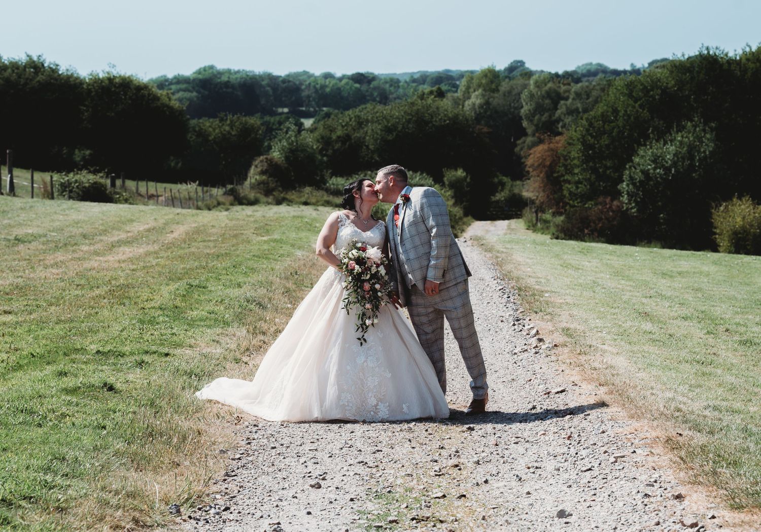 A couple in wedding attire share an embrace on a gravel path surrounded by rolling green countryside and trees.