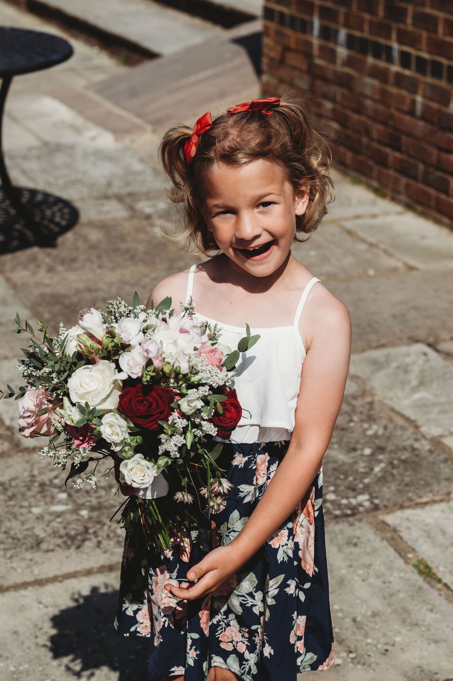 A young person in a white and floral dress holds a beautiful bouquet with red and white flowers against a brick background.