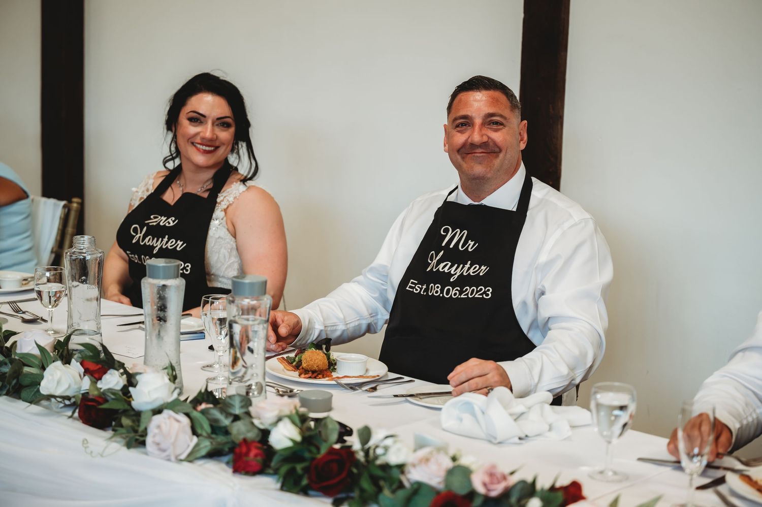 A couple wearing matching Mr and Mrs aprons sit together at their wedding reception table.