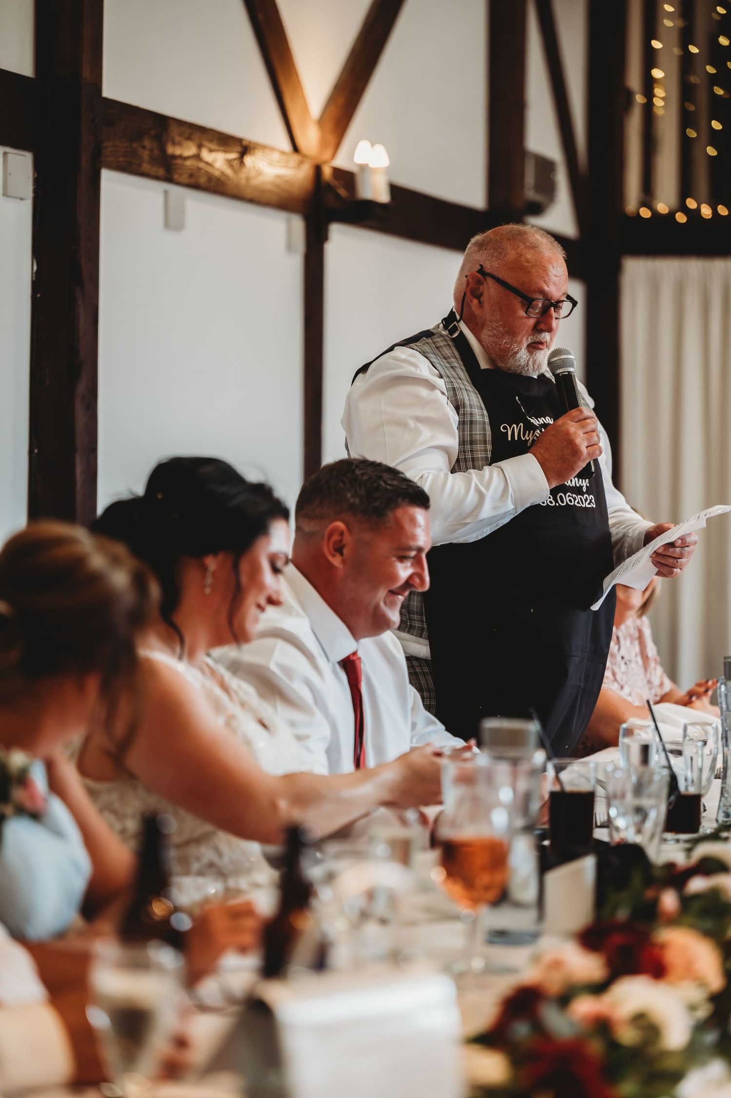At a wedding reception, guests sit at a decorated banquet table while a speaker delivers a toast in a rustic venue.