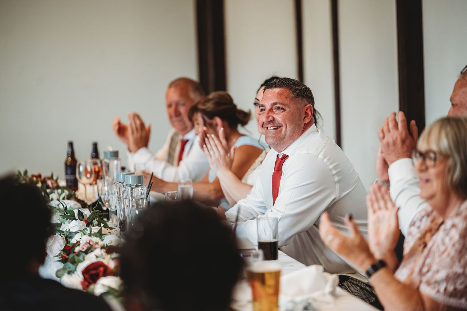 Wedding reception guests in white shirts and red ties laugh and clap during dinner celebration with beer and floral decor.