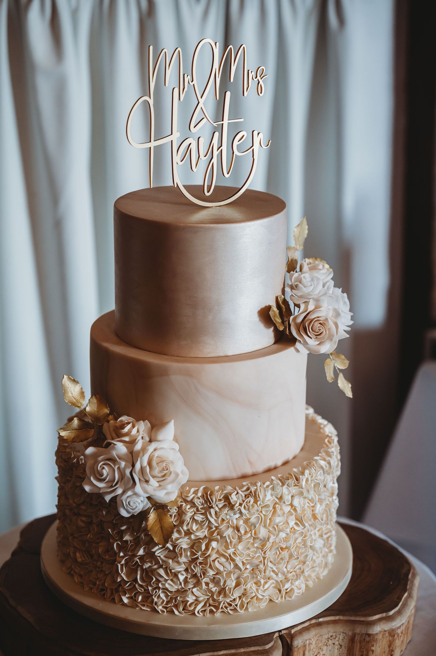 Elegant three-tiered wedding cake with gold accents, textured bottom layer, and delicate rose decorations in cream colors.