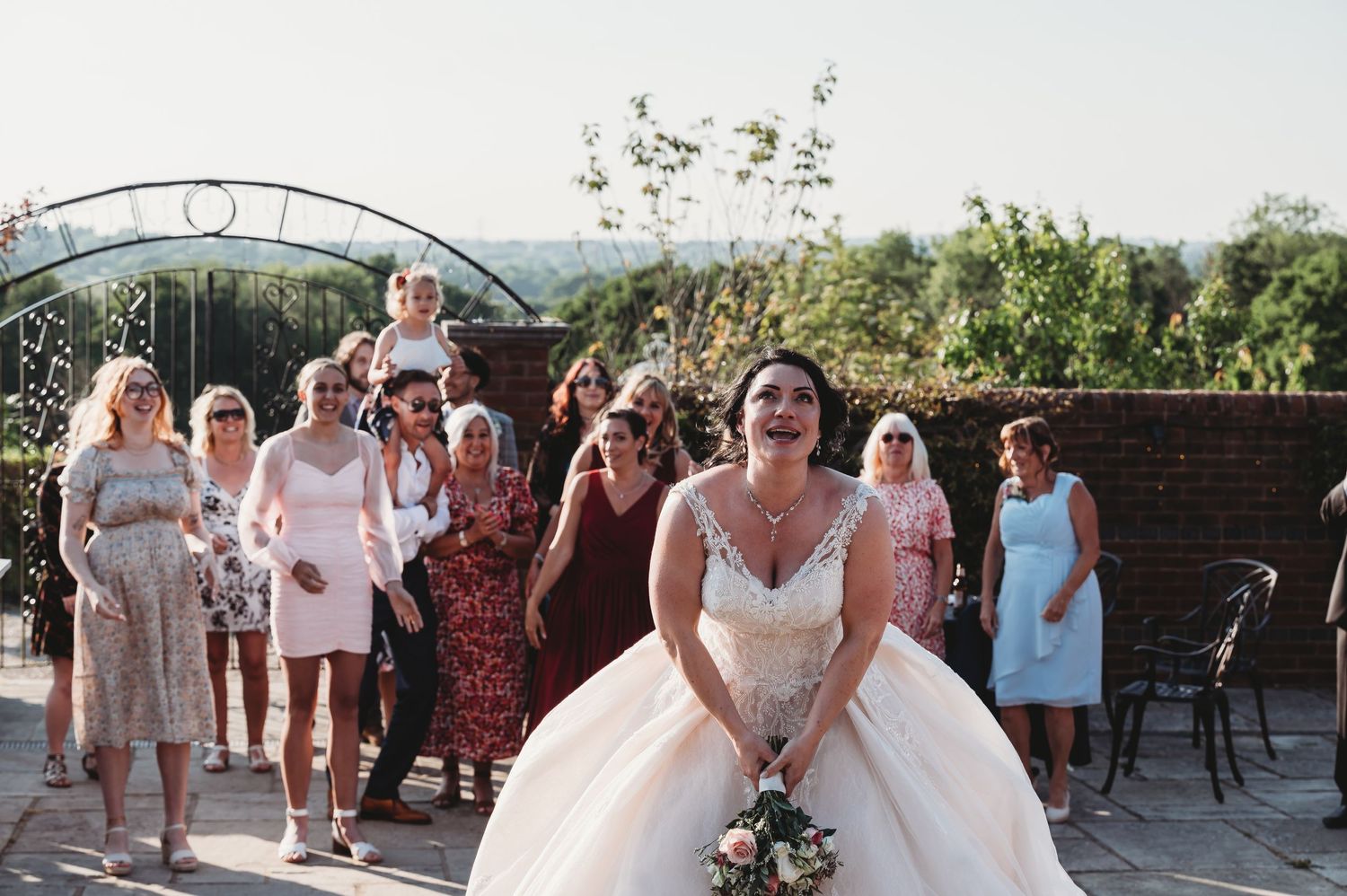 A wedding celebration with guests gathered on an outdoor patio as a bride in a white ballgown poses with her bouquet.