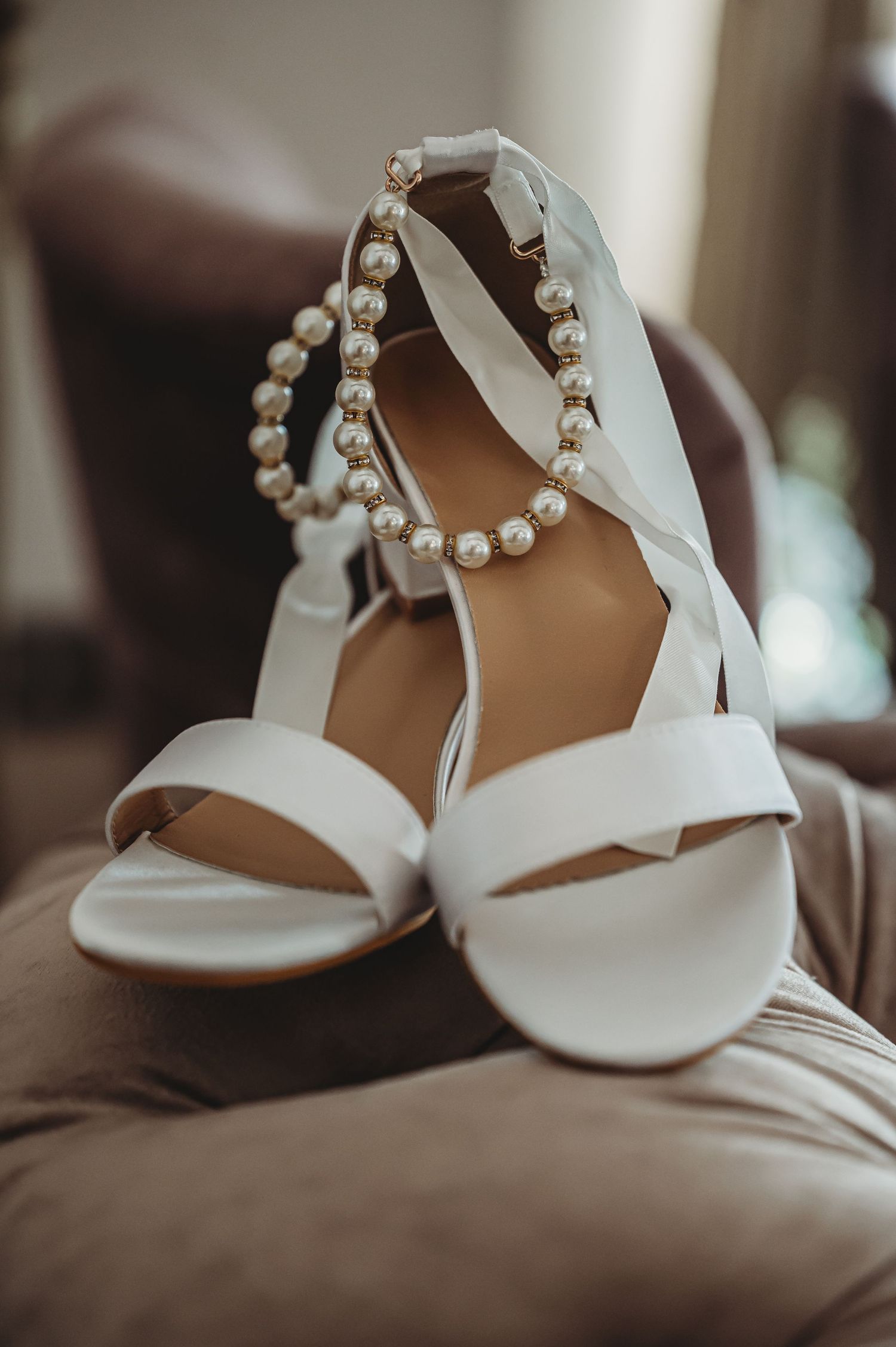 White bridal sandals with ribbon ankle straps displayed alongside a pearl necklace on a brown leather surface.
