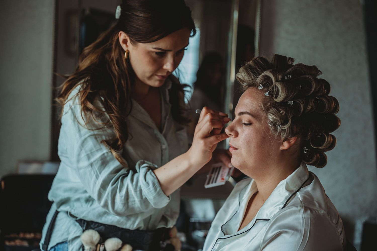 Makeup artist applies cosmetics on client wearing hair rollers in an intimate vintage-styled preparation scene.