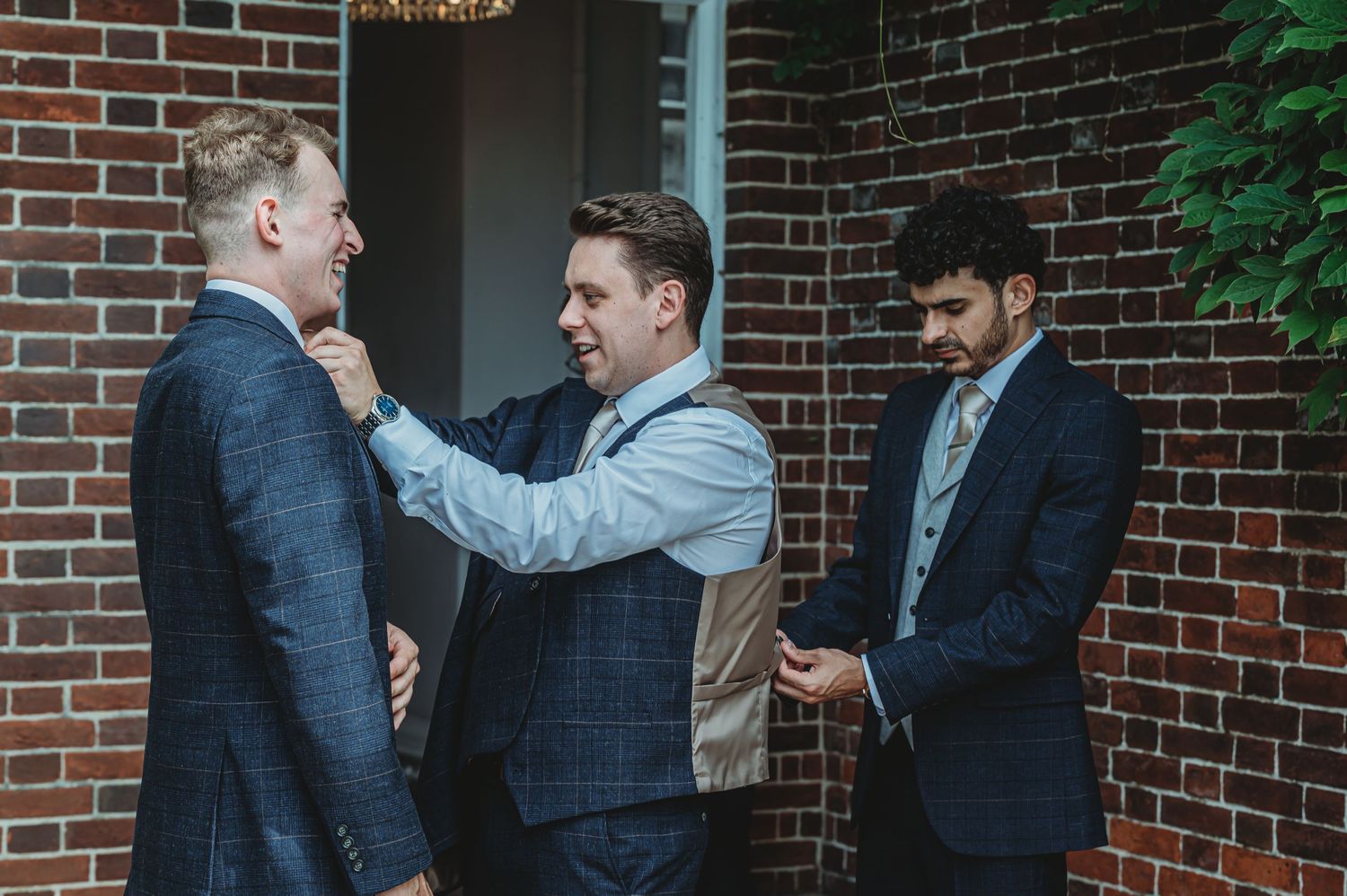 People helping someone adjust their vest and tie while getting ready in front of a brick building.
