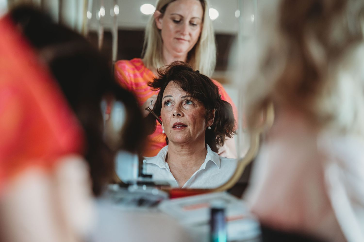 A hairstylist works on a client's hair while both are reflected in a salon mirror.