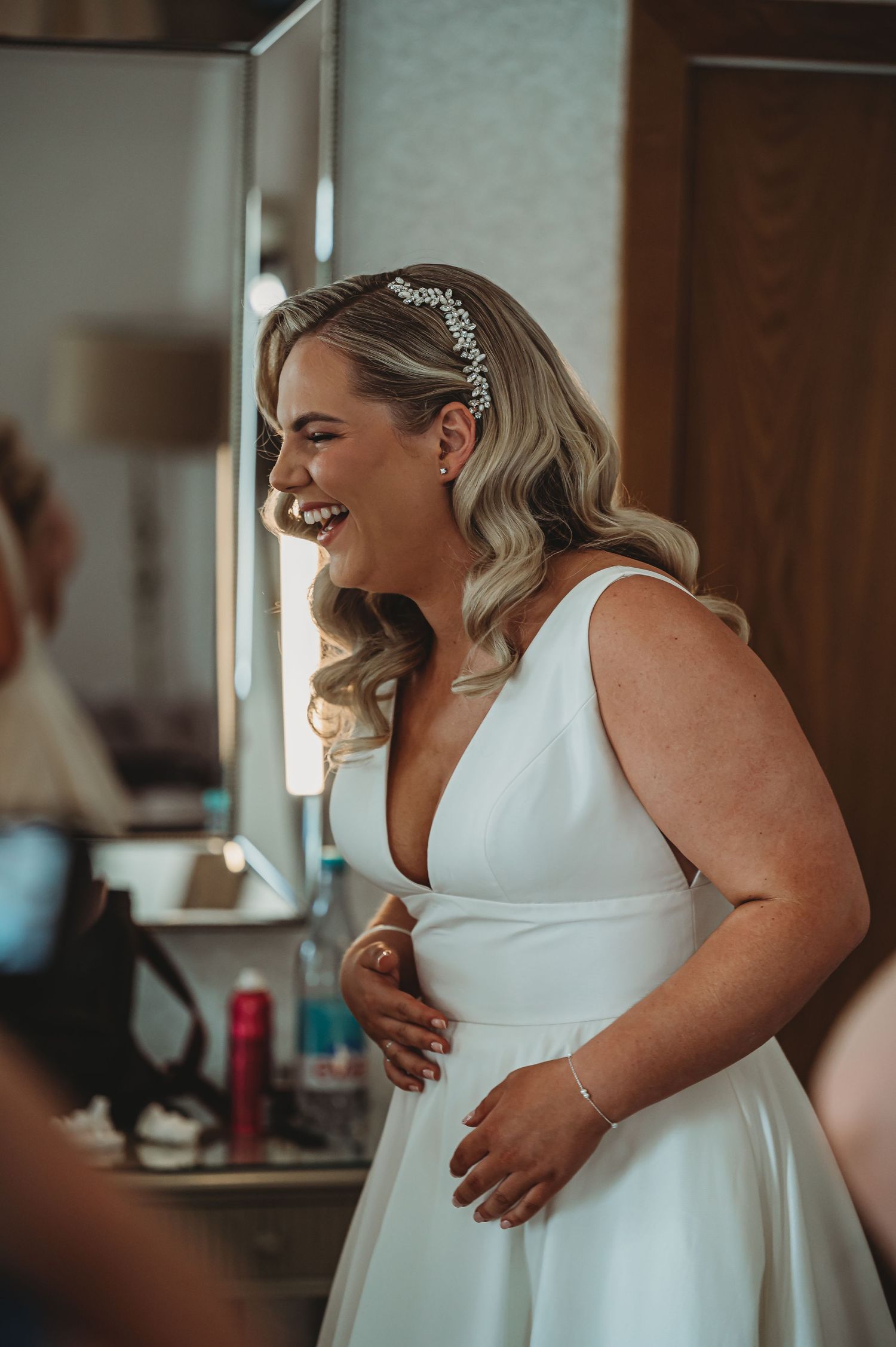 A joyful bride wearing a sleek white wedding dress and crystal headpiece laughs while getting ready for her special day.