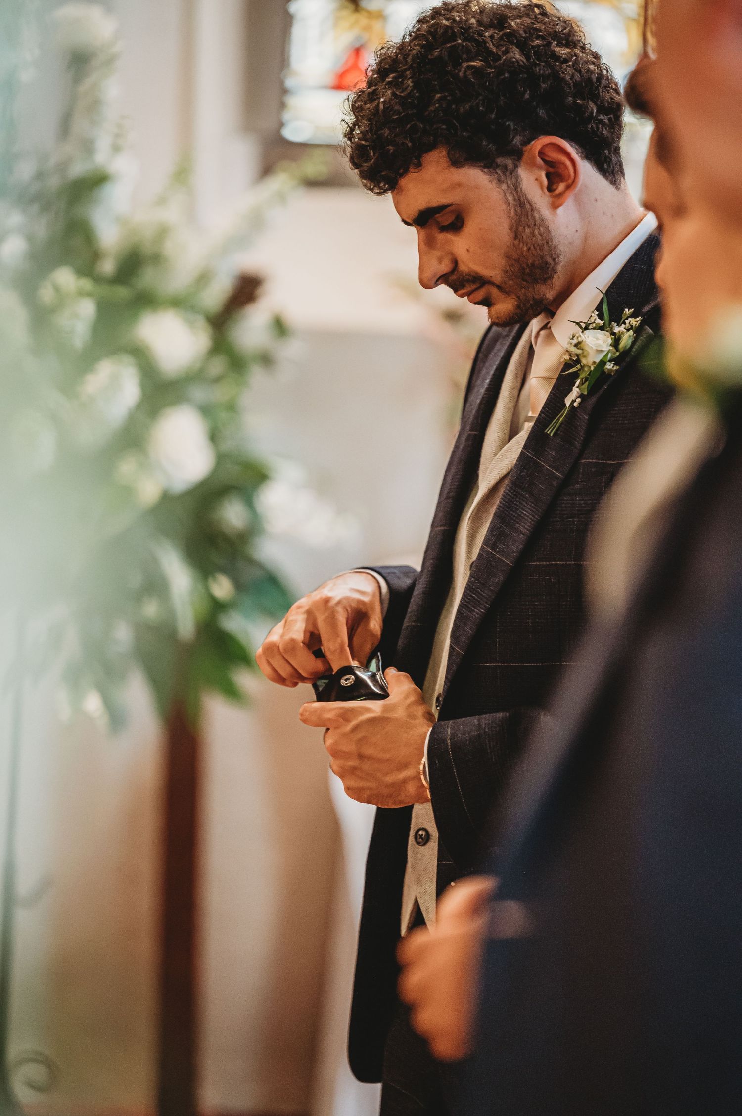 A groom wearing a dark suit with boutonniere checks his phone during a quiet moment at the wedding ceremony.