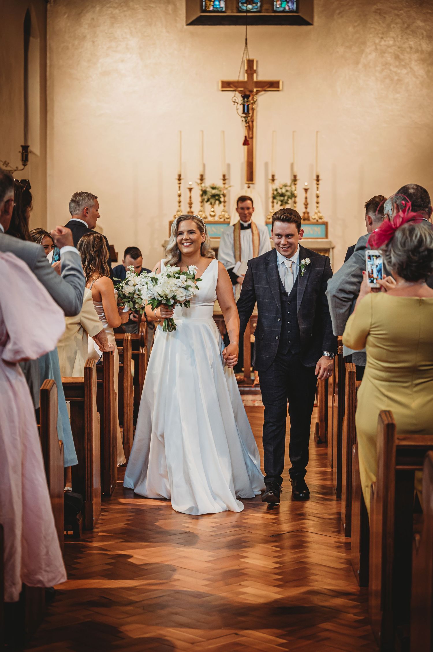 A bride and groom walk down a church aisle with wooden pews and a crucifix on the wall during their wedding ceremony.
