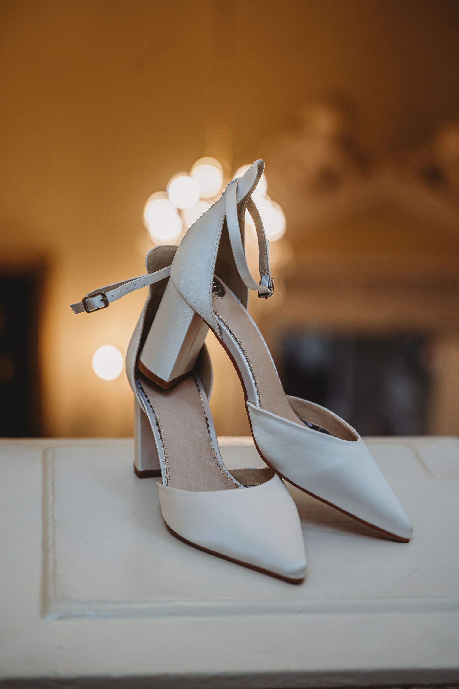 Elegant white bridal heels with pointed toes and ankle straps sit gracefully against a warm bokeh background.