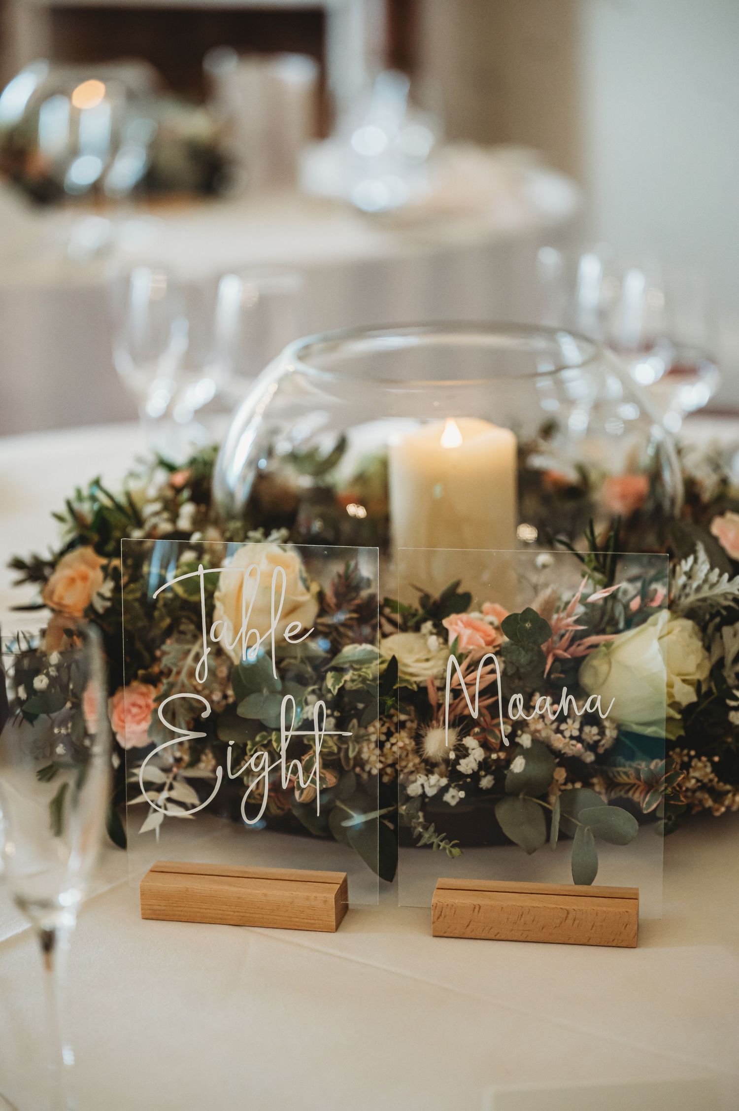 Elegant wedding table number display with clear acrylic signs, candles, and peach roses surrounded by greenery centerpiece.