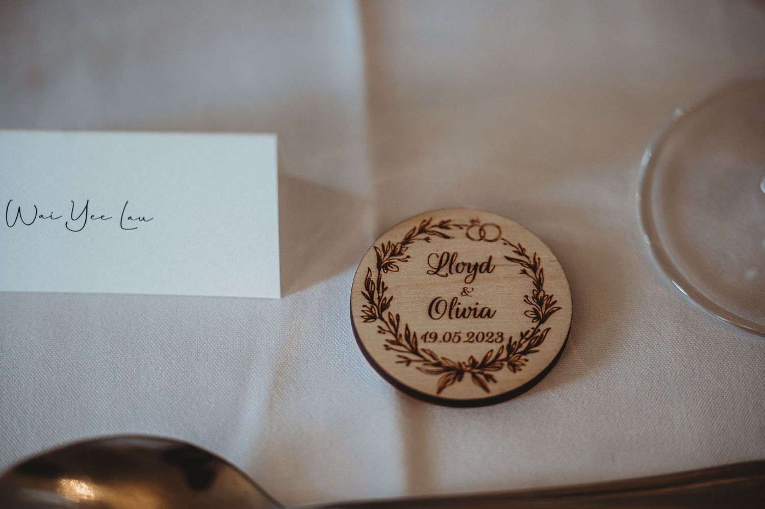 A wooden round place card with an elegant wreath design and names engraved sits on a white tablecloth.