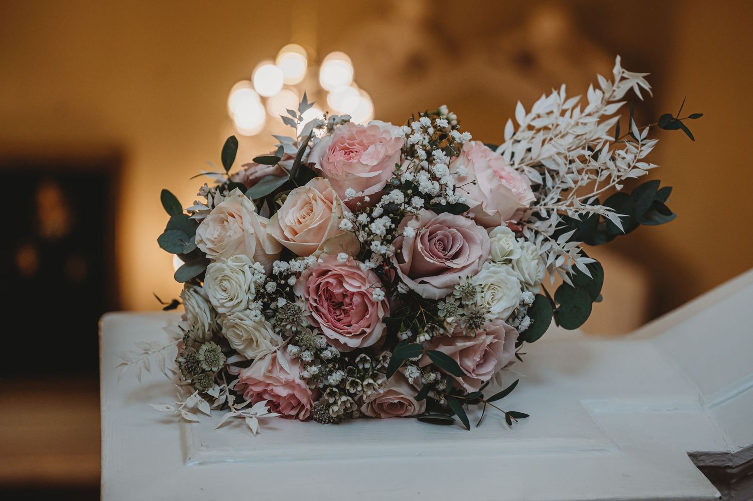 A romantic wedding bouquet with soft pink roses and delicate greenery rests on a white surface in warm lighting.