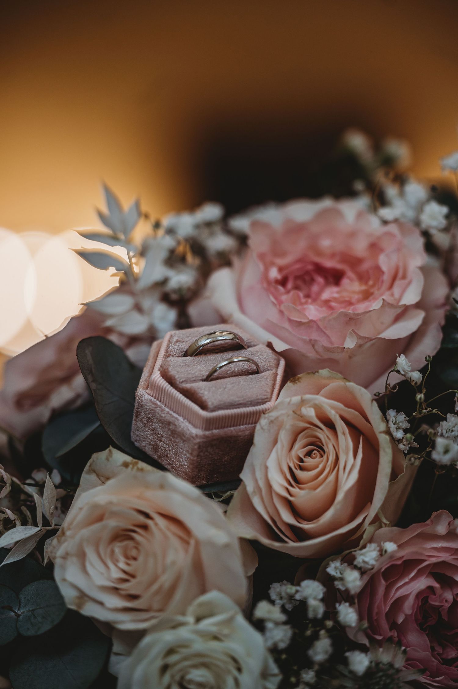 Close-up photo of wedding rings nestled among pink and cream roses with baby's breath flowers in soft lighting.
