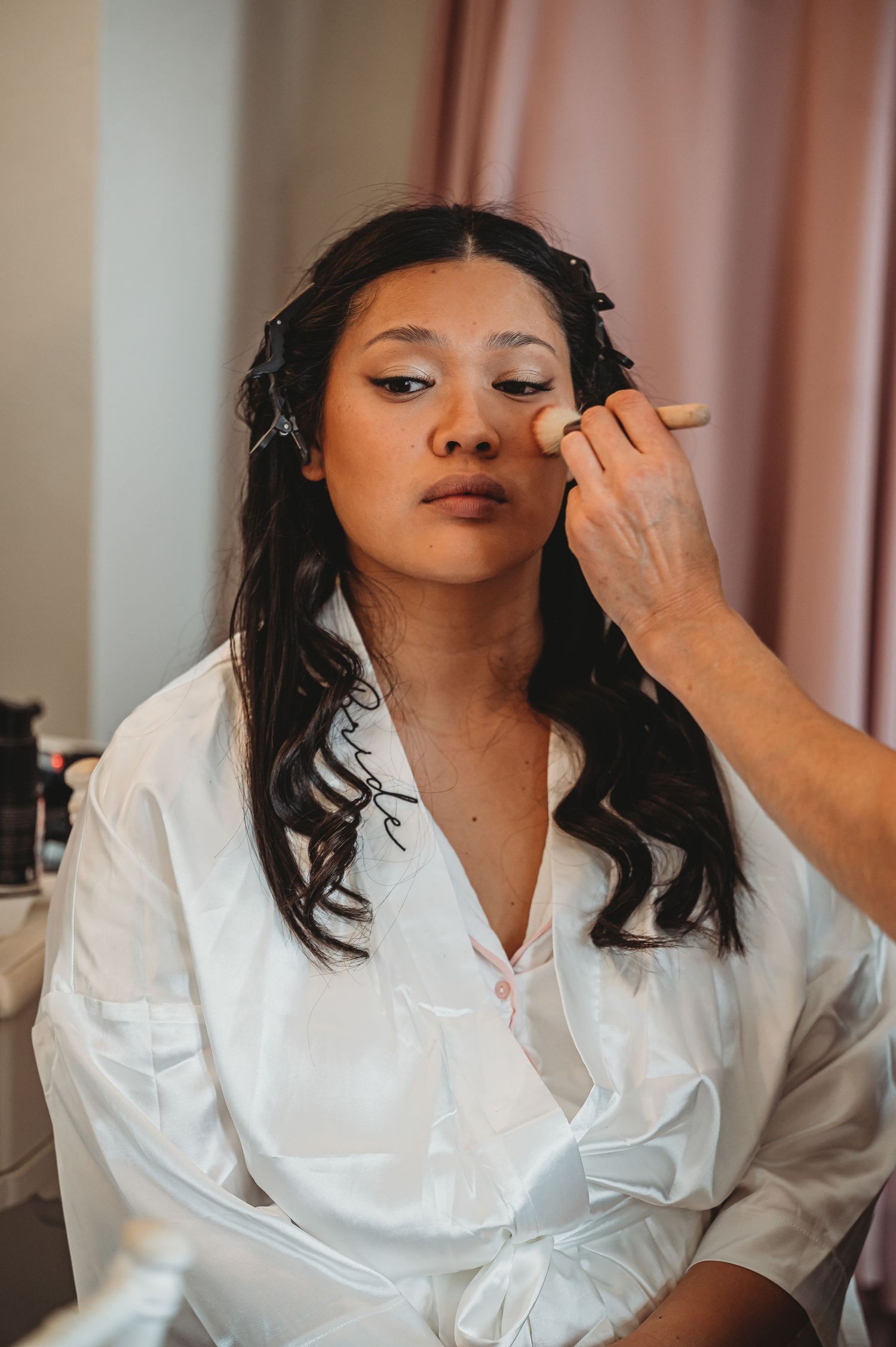 A makeup artist applies cosmetics to a woman wearing a white silk robe during a beauty preparation session.