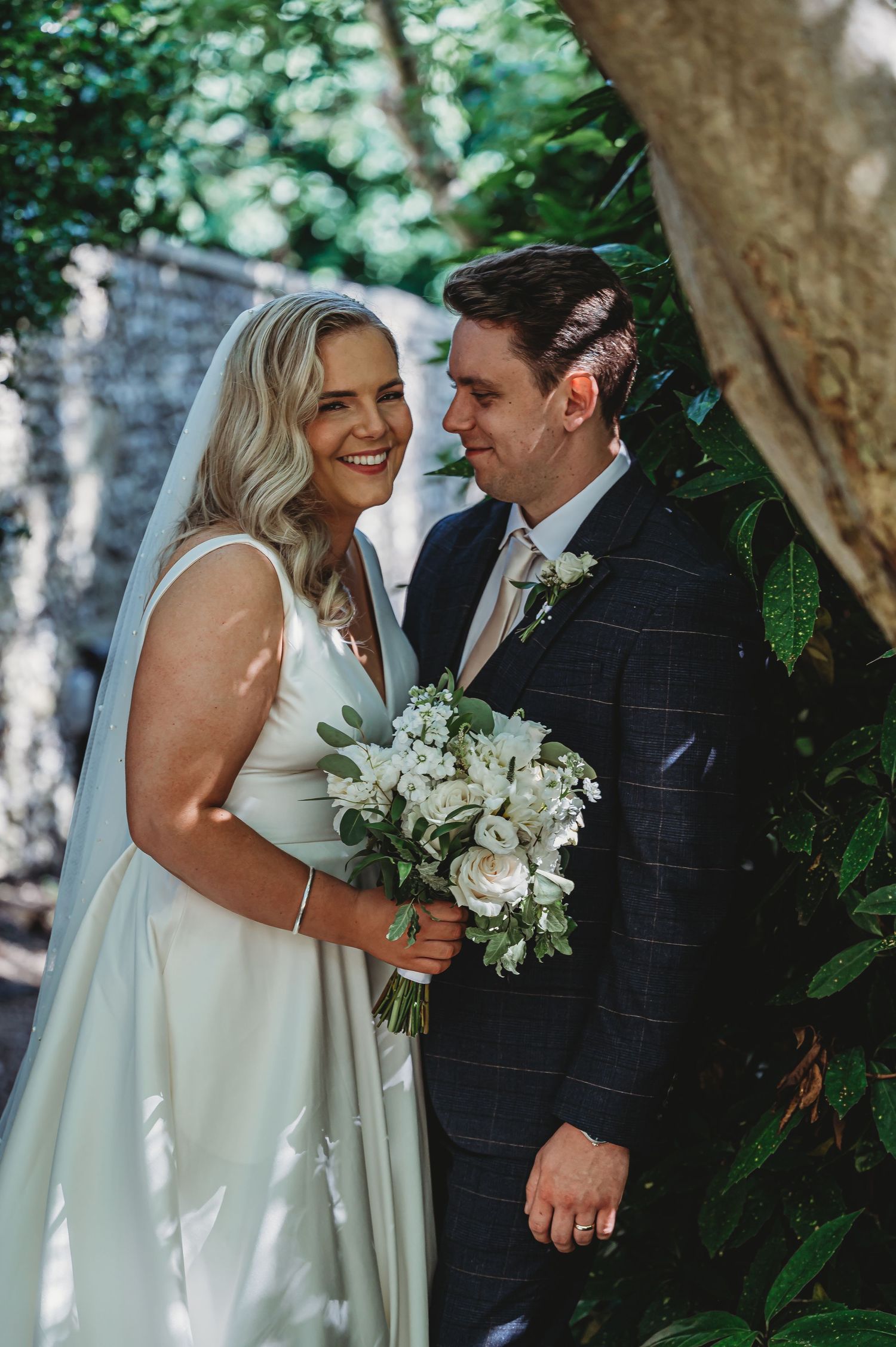 Outdoor wedding portrait with blooming greenery backdrop showing happy couple with white flowers in dappled sunlight.