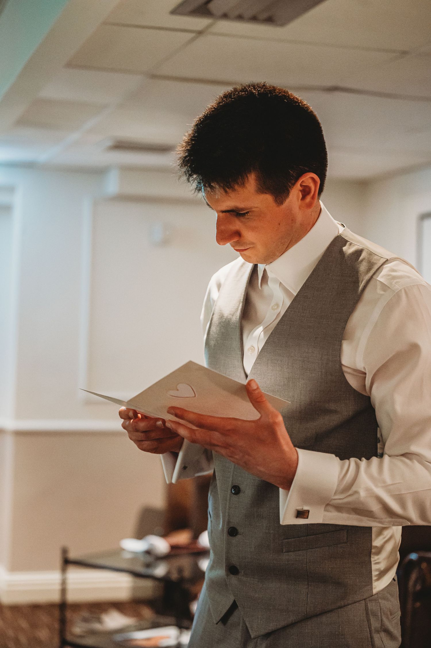 A groom in a grey vest and white shirt reads a paper document while standing in an indoor room.