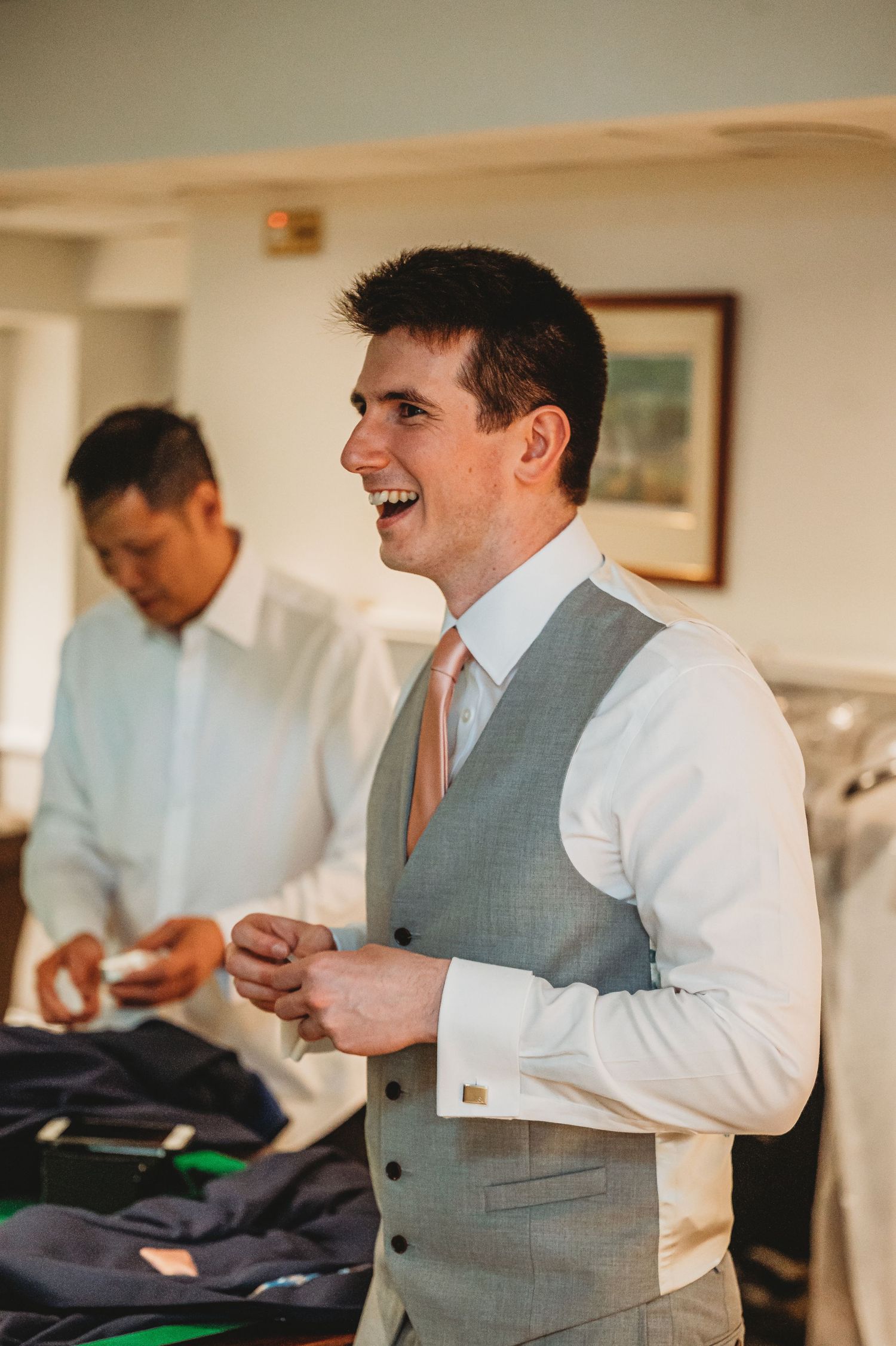 Joyful groomsmen share a laugh while getting ready on wedding day, wearing formal attire with grey vest and peach tie.
