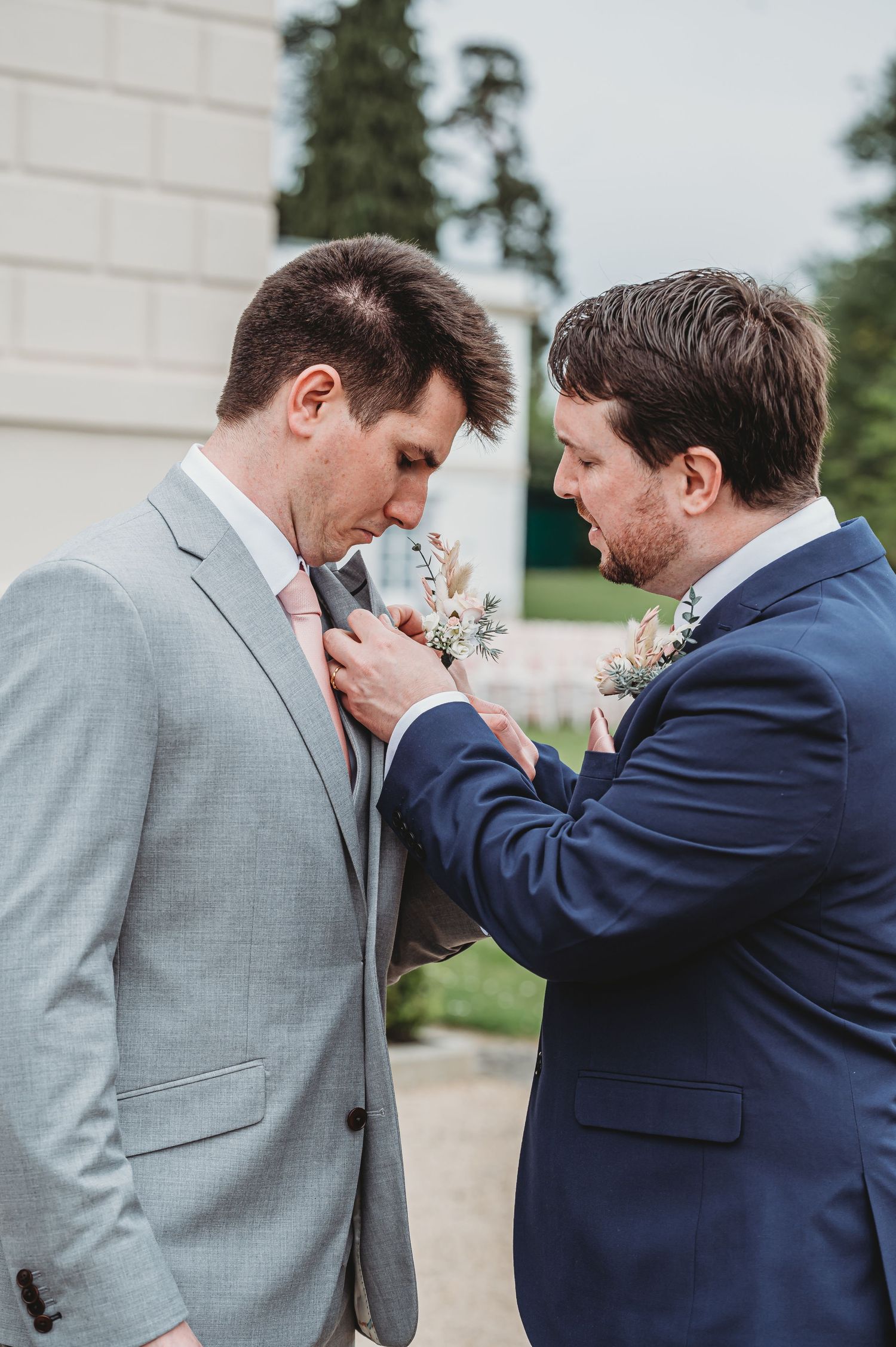 Two people in formal suits helping each other with boutonniere flowers during a wedding ceremony outdoors.