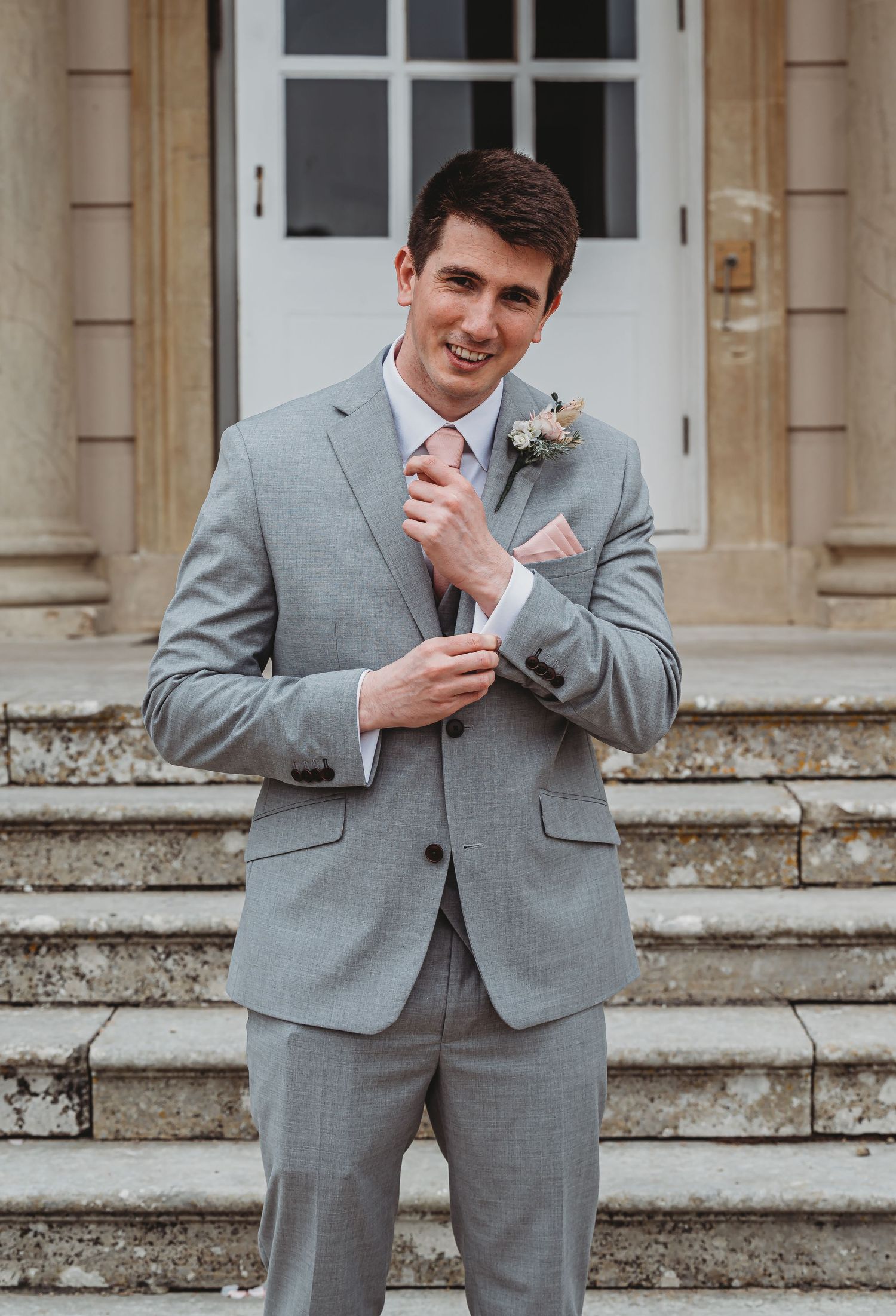 A groom in a light gray suit with pink tie and boutonniere stands on stone steps outside a building.