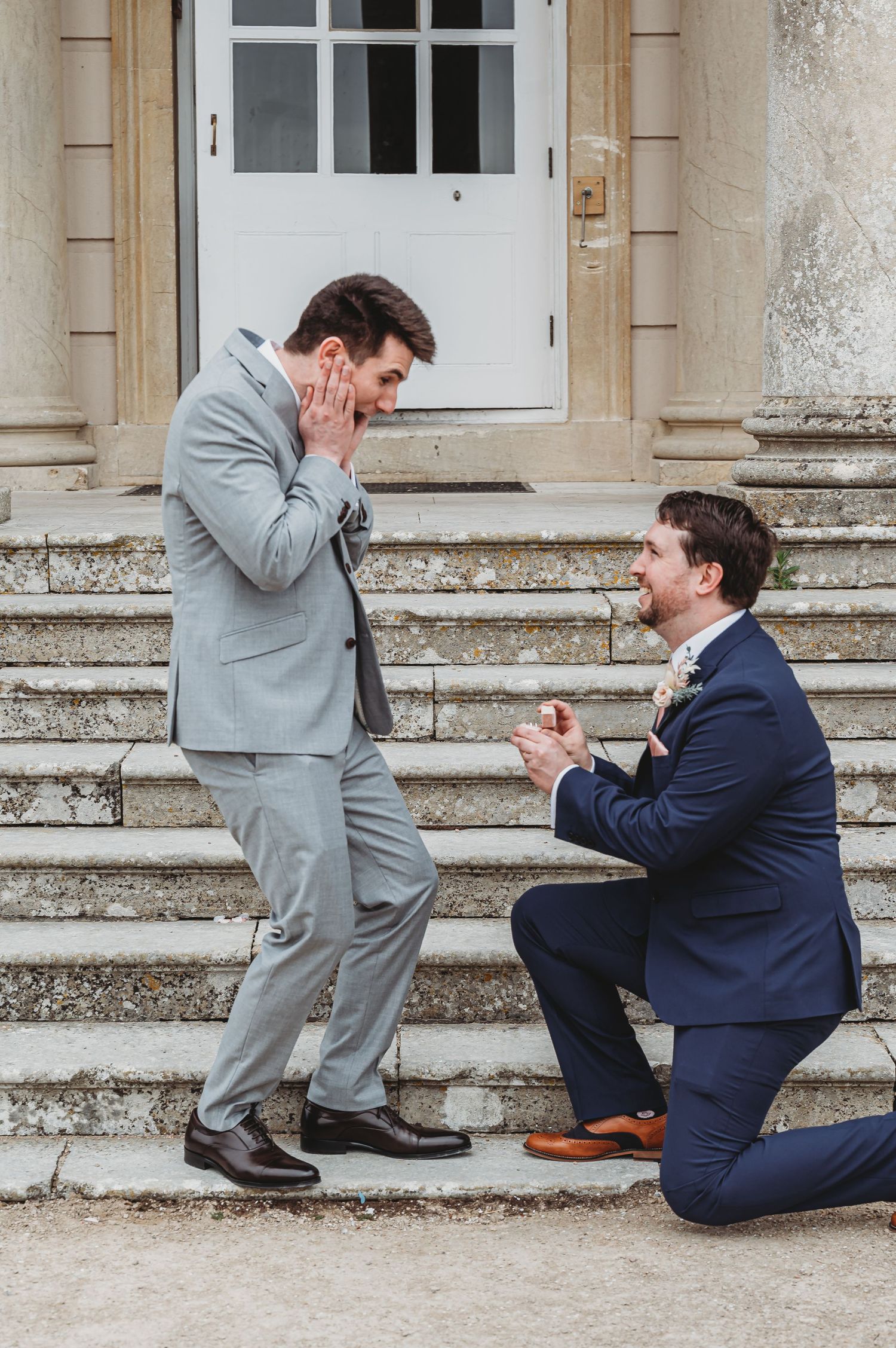 A surprise marriage proposal taking place on stone steps outside a building with one person kneeling in a navy suit.