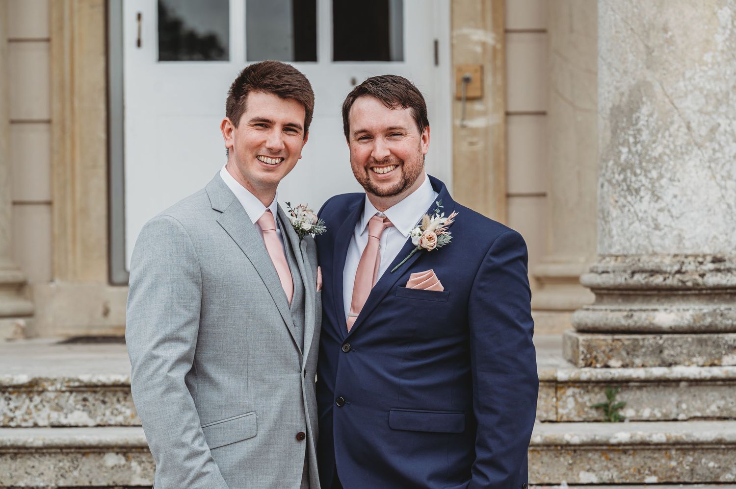 Two grooms wearing coordinated suits in light grey and navy blue with matching boutonnières stand together outside a stone building.