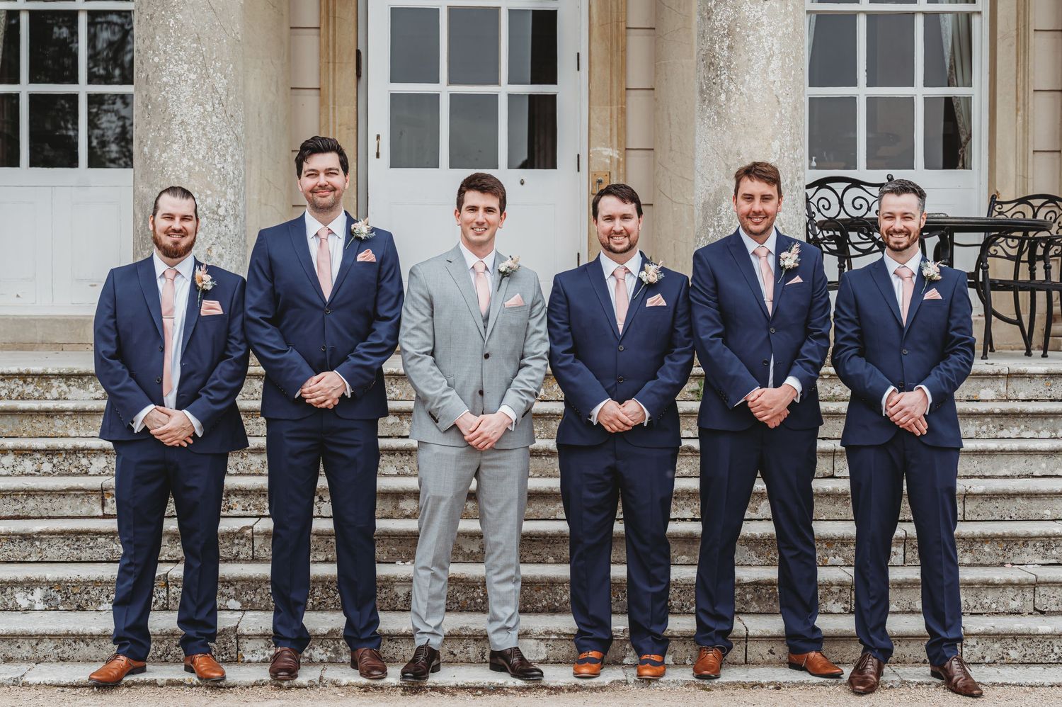 A group of groomsmen in navy and gray suits stand lined up on stone steps in front of a traditional building.