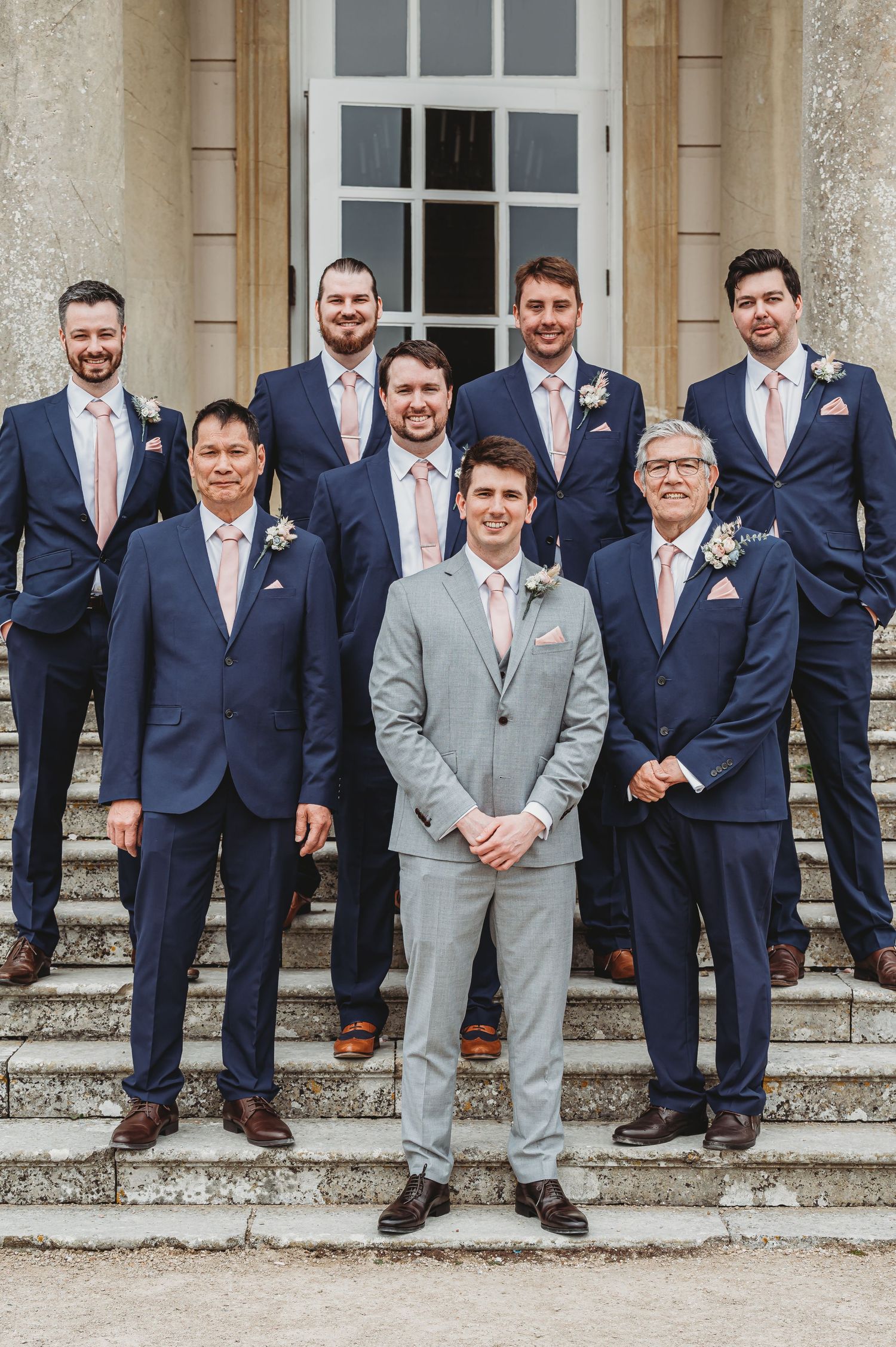 Groomsmen in coordinated navy suits and one in light gray stand on stone steps outside an elegant building for a wedding photo.