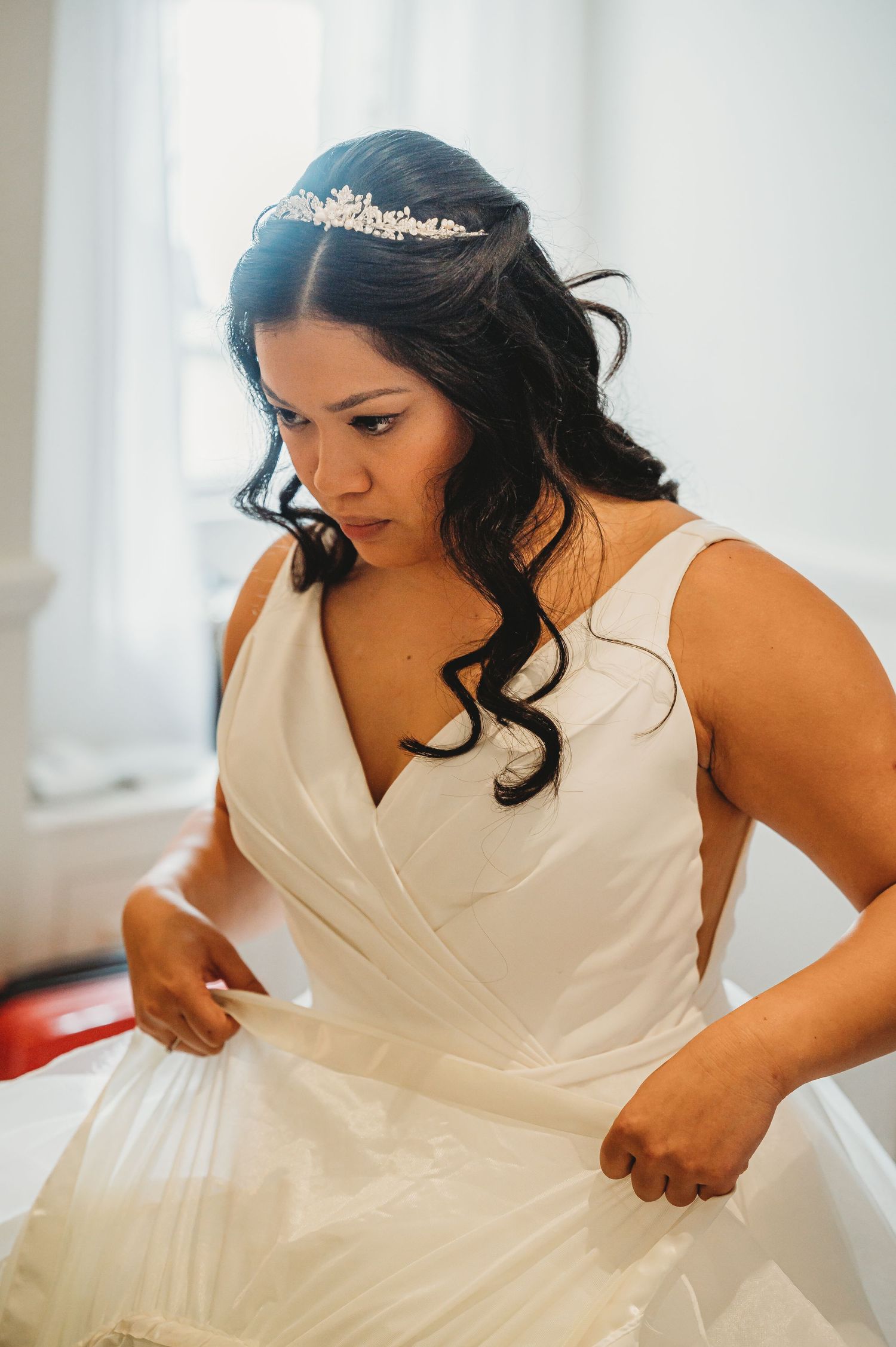 A bride in an elegant white V-neck wedding dress adjusts her gown while getting ready, wearing a delicate crystal tiara.