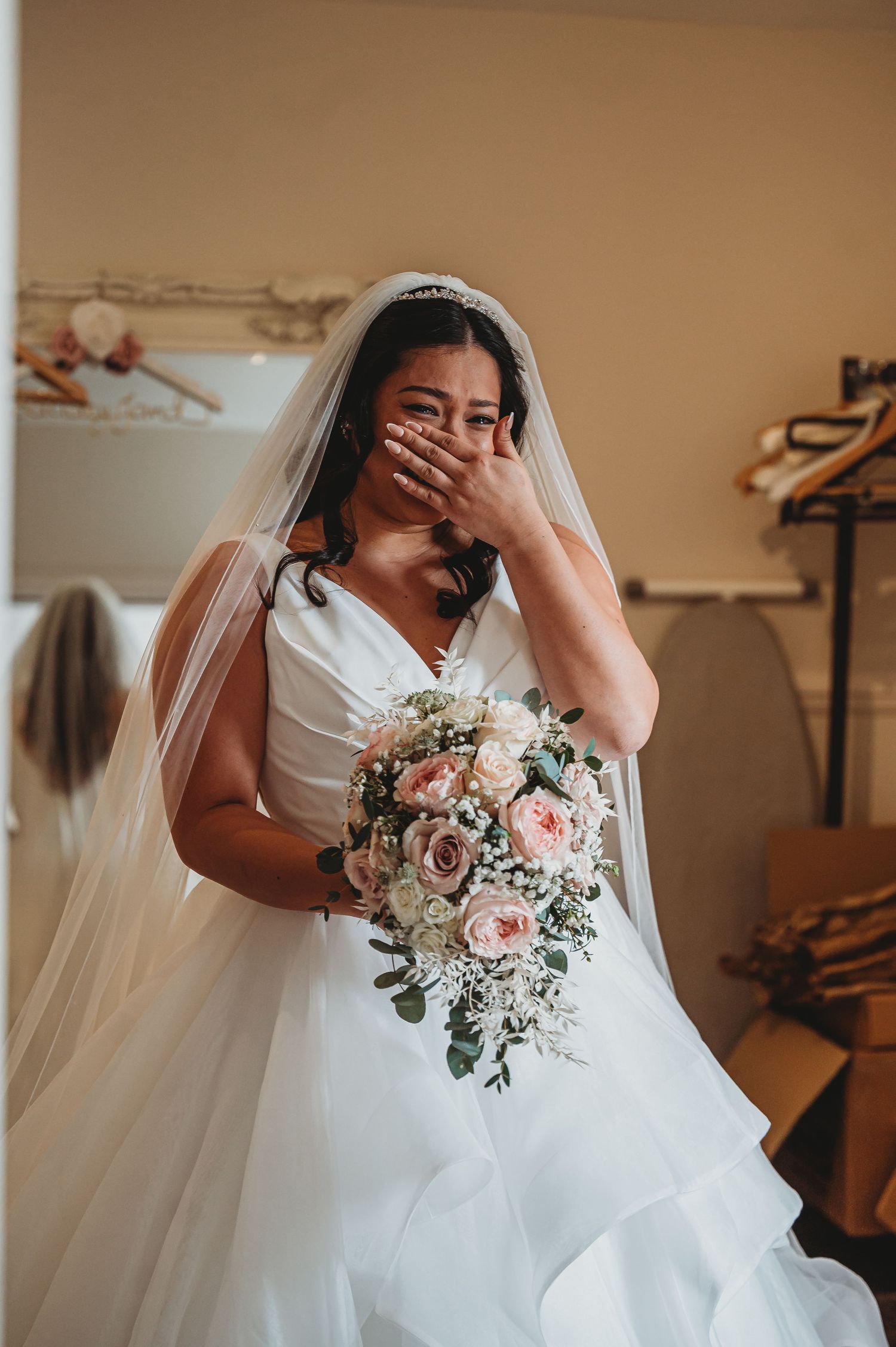 A beautiful bride in a white gown and veil holds a cascading pink and white bouquet while covering her face in joy.