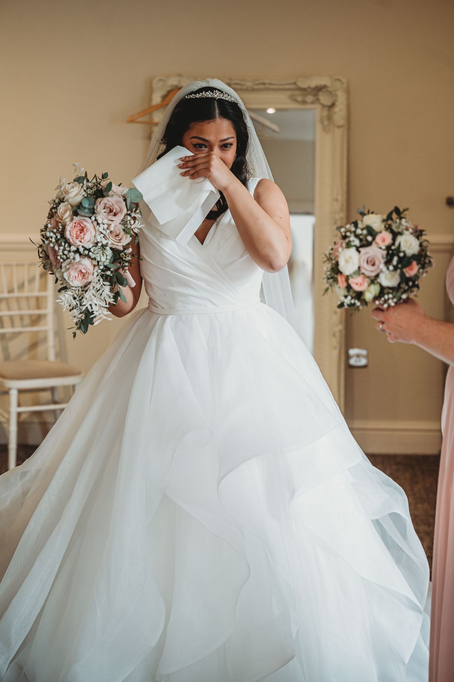 A bride in a flowing white ballgown wedding dress dabs her eyes with a tissue while holding a pink and white bouquet.