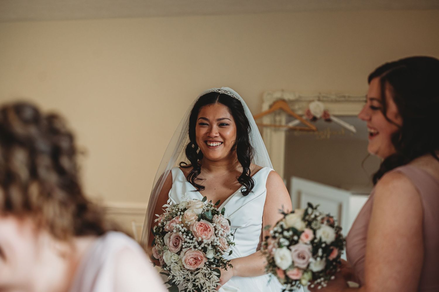 A bride in a white dress with floral details smiles while holding her bouquet during wedding preparations.