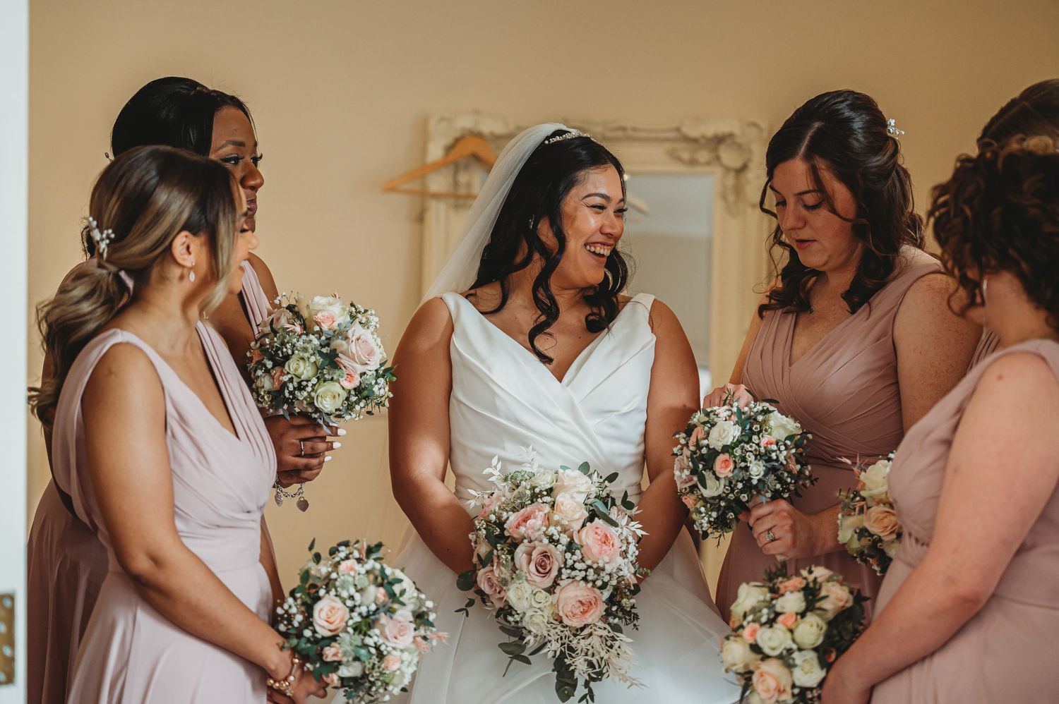 A group of women in blush pink and white dresses hold pastel floral bouquets while sharing a joyful moment together.