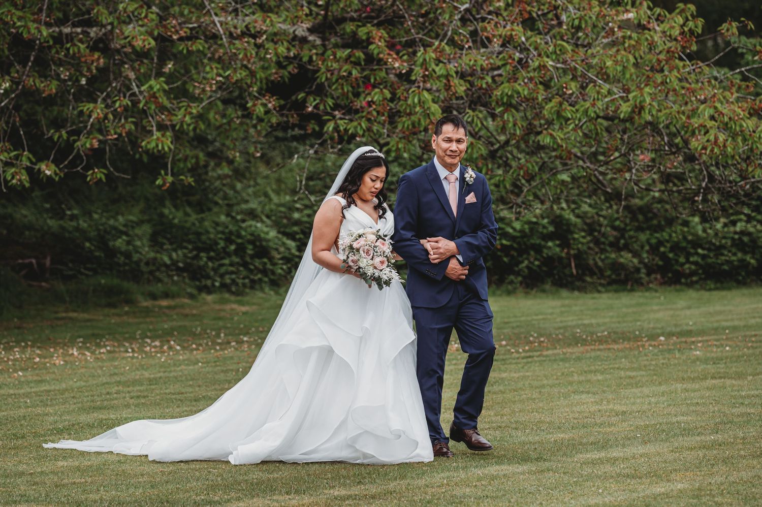 A bride in a flowing white gown and groom in navy suit walk together on lush green lawn during outdoor wedding ceremony.
