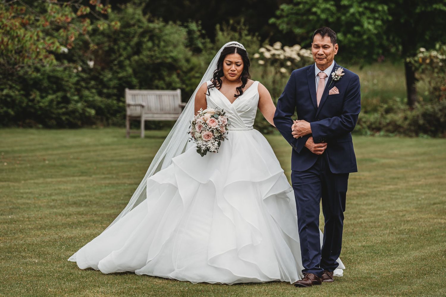 A beautiful outdoor wedding ceremony with a bride in a flowing white gown and groom in navy suit walking on grass.